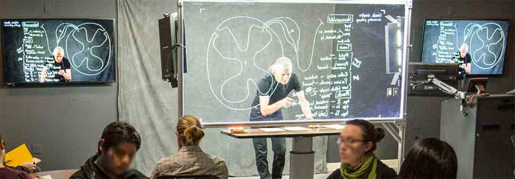 A man stands in front of a large glass board working on the board. Strong lights shine on him from above while his image is reflected in monitors around the classroom