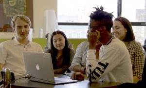 Students gather around a laptop computer listening to a podcast