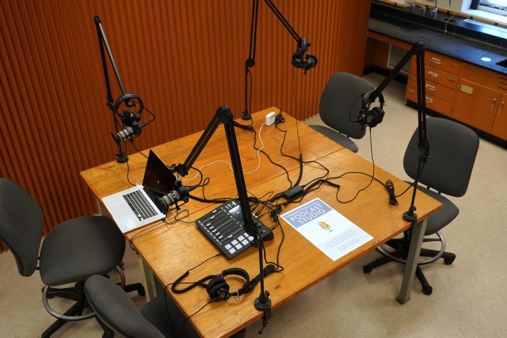 A brown table with 4 microphones and headphones on articulating arms, a laptop and audio mixer sit on the table. A display sign announces "Podcast Studio".