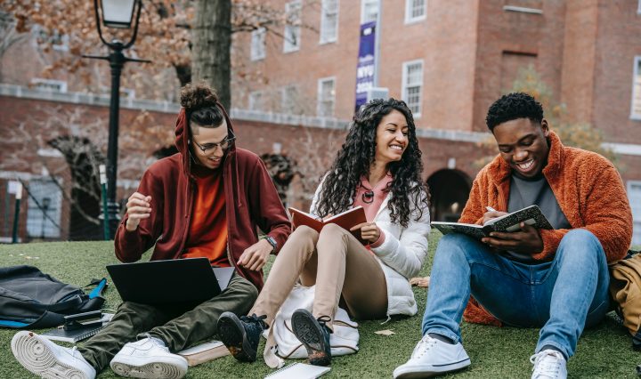 3 students sitting on college campus lawn smiling, reading books, and looking at their laptops.