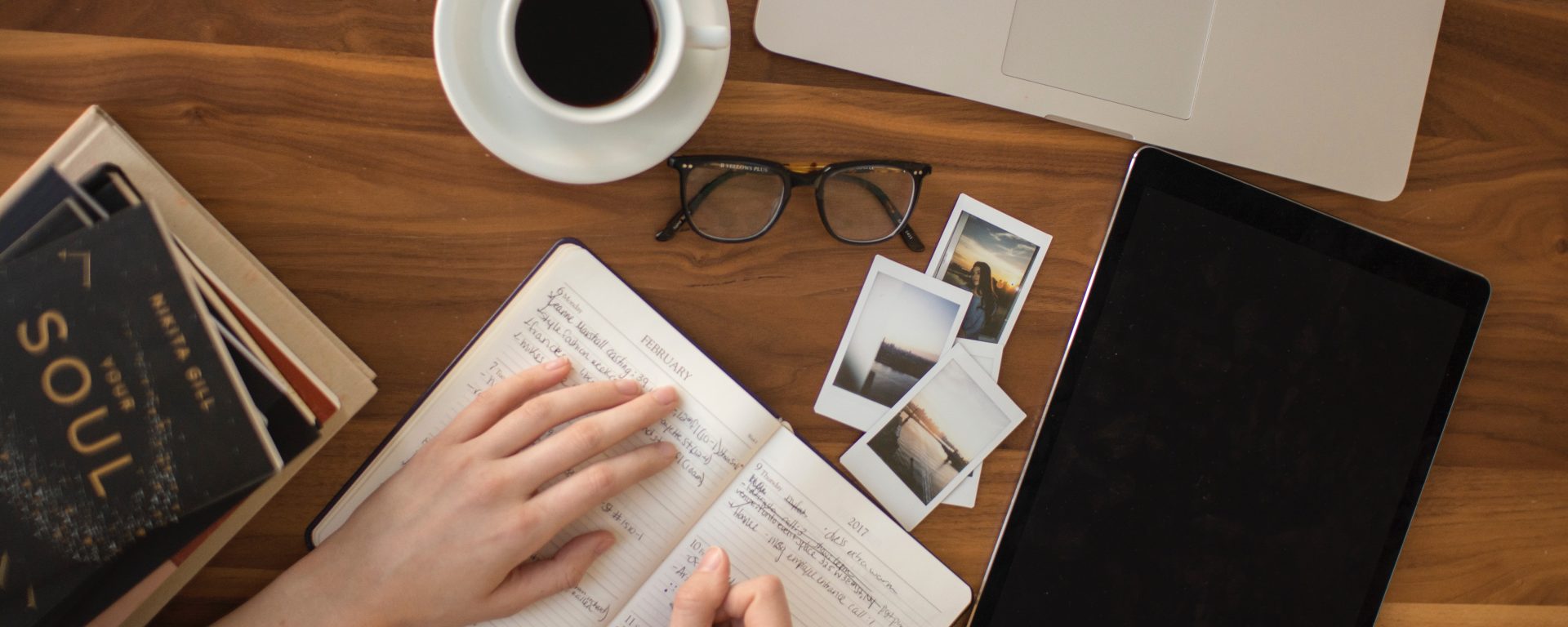 person holding ballpoint pen writing in a notebook with a cup of coffee, eye glasses, and a laptop on a desk.