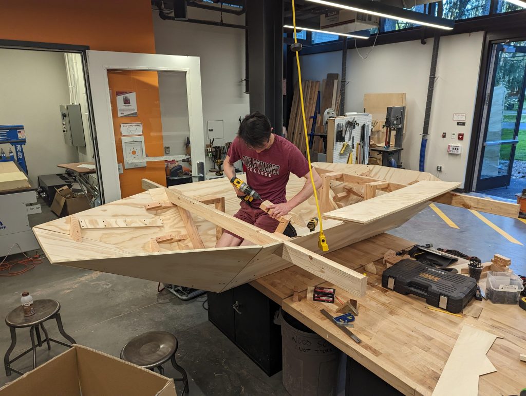 a student inside a plywood boat during construction, on top of a work table in the makerspace