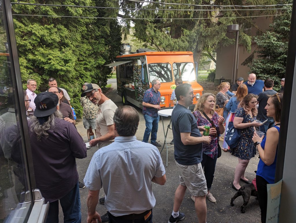 Exterior picture showing a few dozen faculty and staff mingling under decorations and a beside a food truck.