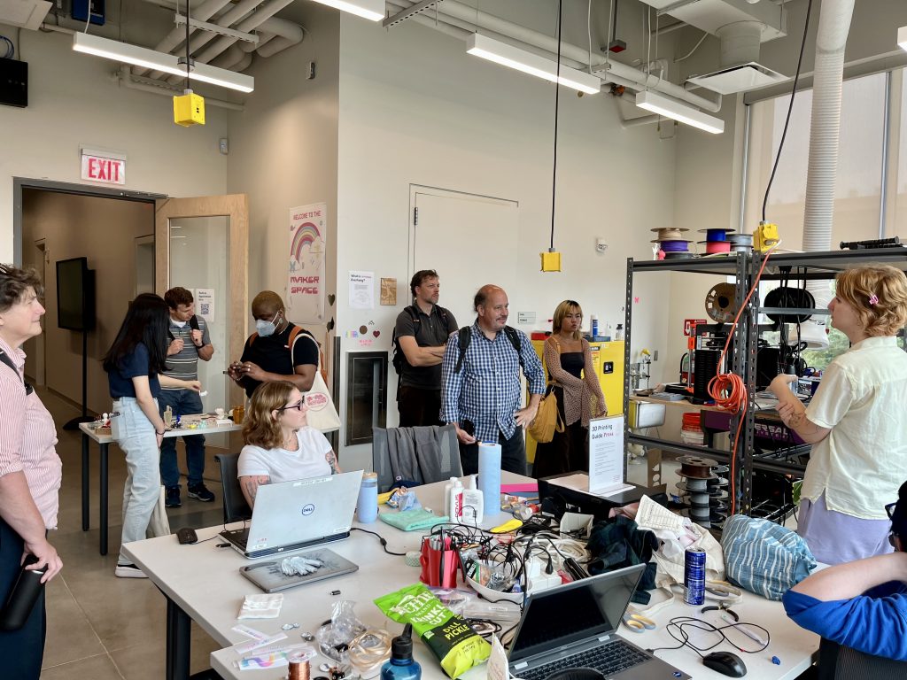 Temple Makerspace staff shows Swarthmore Accessibility team members around the Makerspace. There are electrical boxes hanging from the lofted ceiling; desks with various devices and materials; shelving units with 3D printers on them.