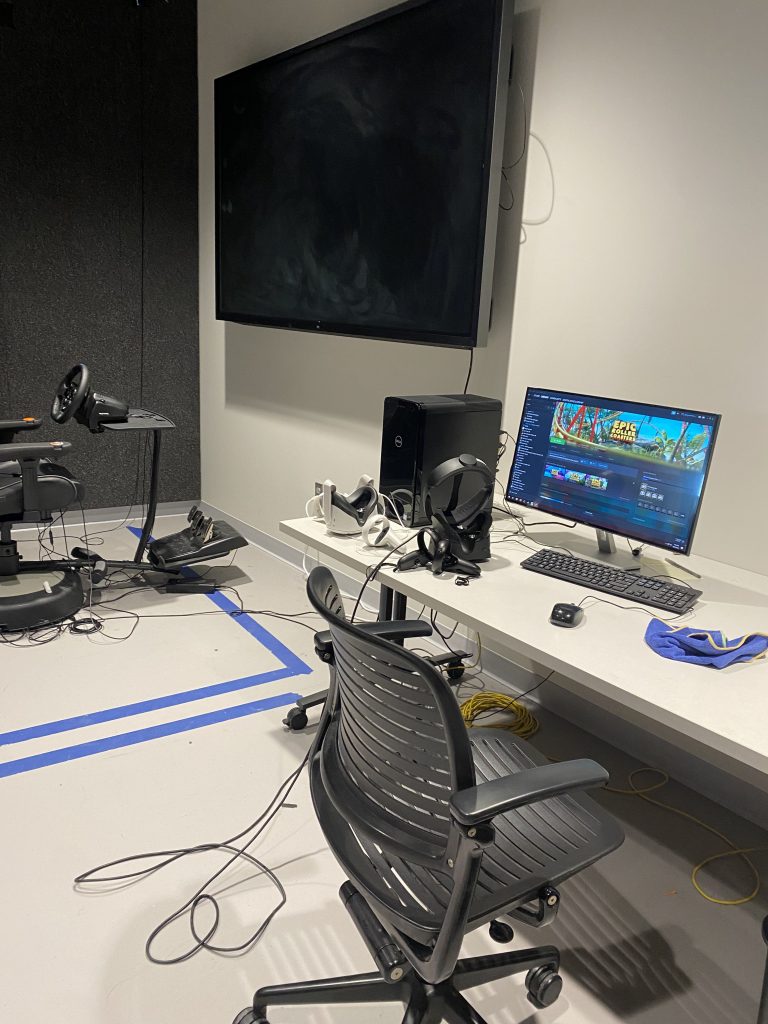 VR studio equipped with multiple VR headsets and large screens. There is painter's tape on the floor to map out space for each user. A driving simulator can be seen in the background.