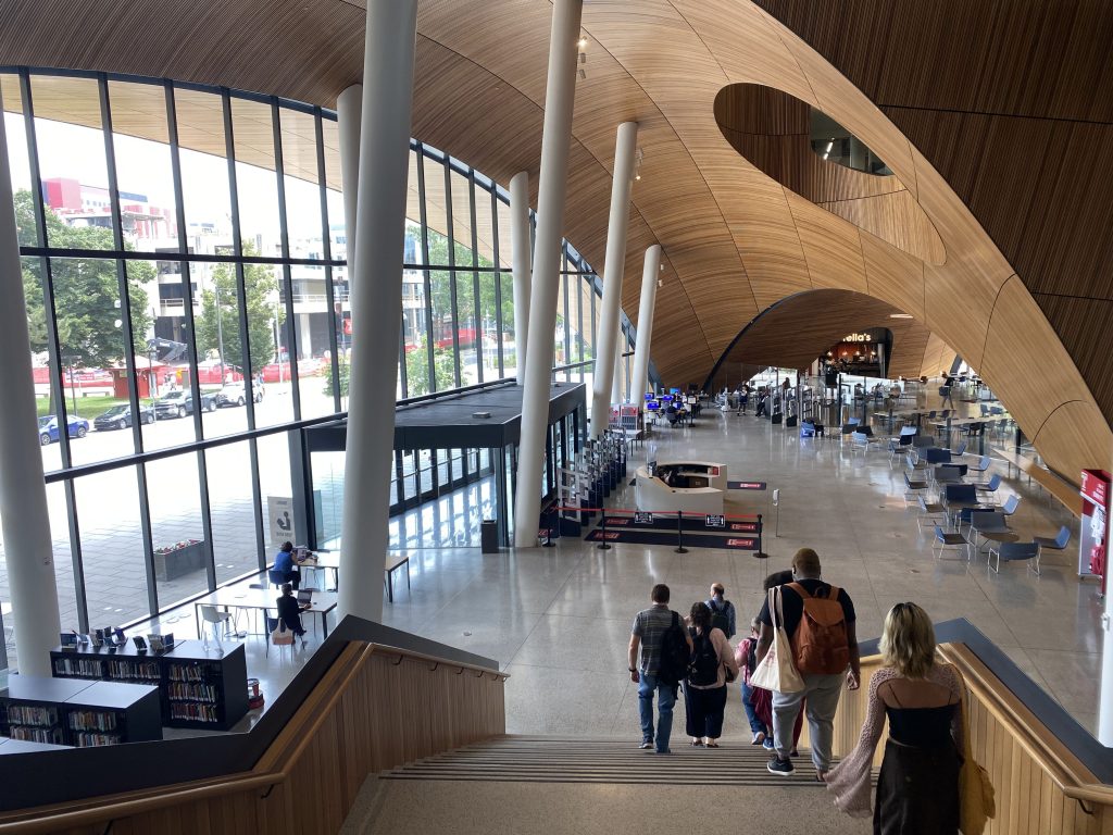 View of the first floor of Charles Library from the atrium stairs. Ceiling-to-floor length windows are on the left with wood in a curved pattern making up the ceiling and opposite walls, with an archway opening to the cafe on the opposite side from the picture.