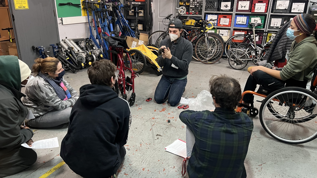 TechOWL employee demonstrates a type of Assistive Technology to workshop attendees. They are surrounded by various mobility devices, and the people are sitting on the ground.