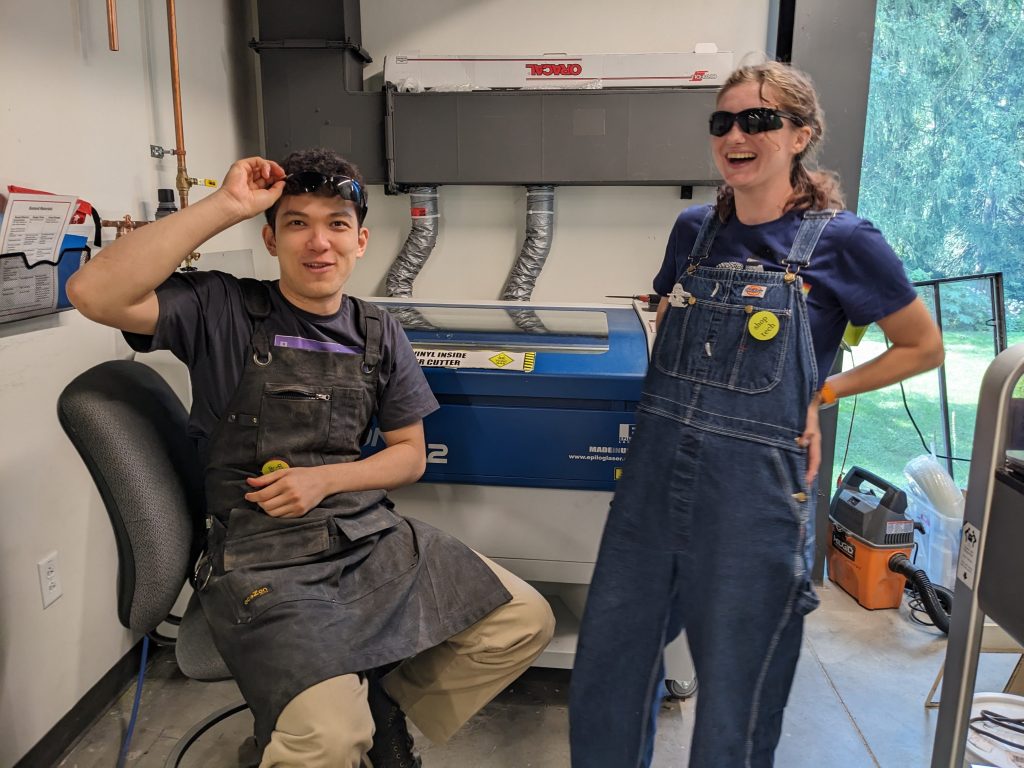 Students Miranda and Myles laugh posing for the camera in the laser cutter room
