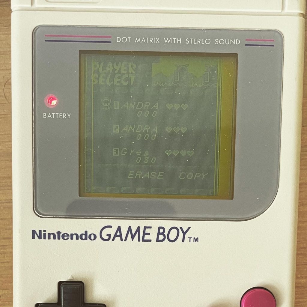 Front view of an original Nintendo Game Boy with a game screen showing 'PLAYER SELECT' and a low battery indicator, on a wooden background.
