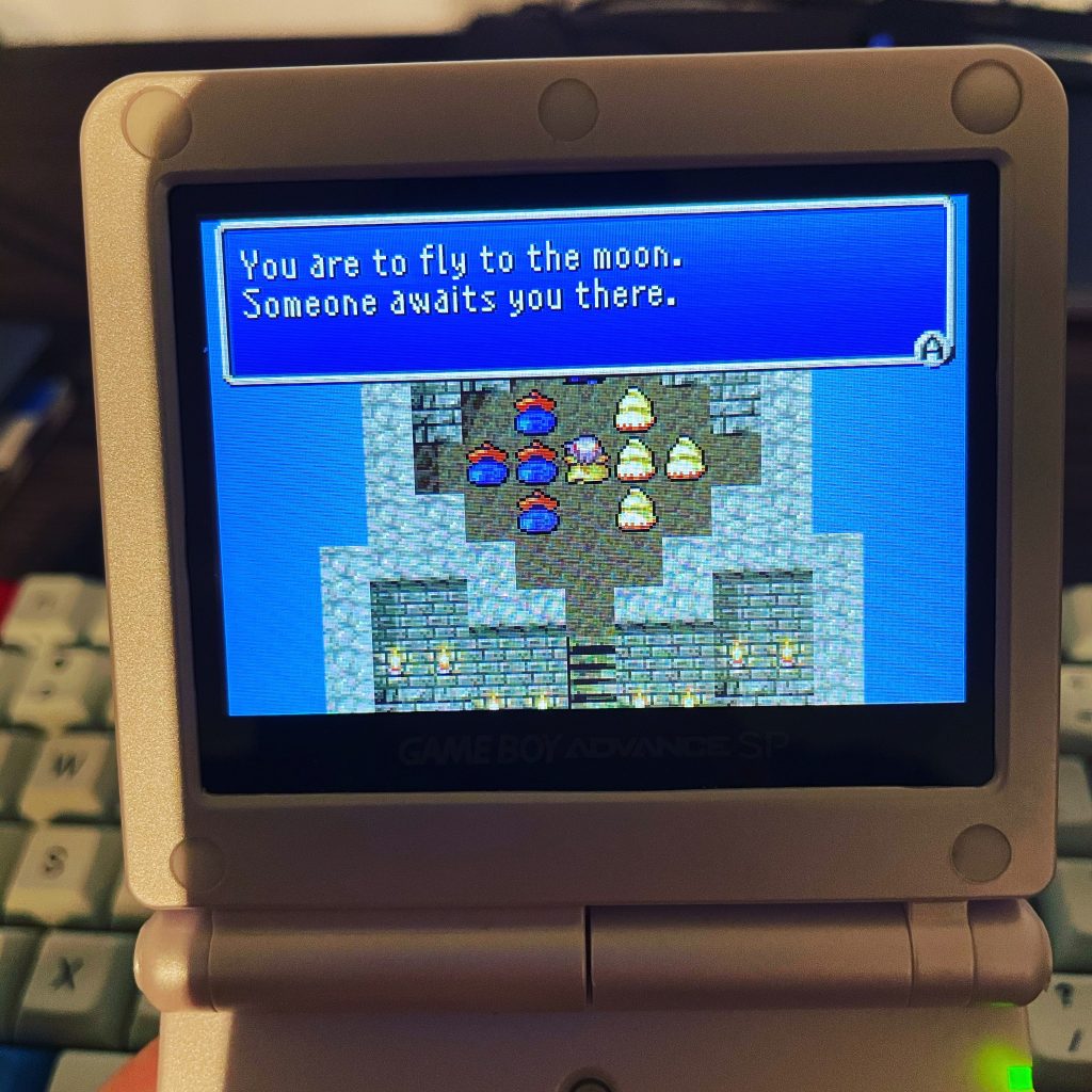 Close-up of a silver Game Boy Advance SP with a game screen displaying dialogue 'You are to fly to the moon. Someone awaits you there.' with pixelated characters, held in hands with a keyboard in the background."