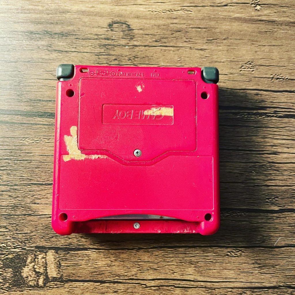 Top view of a worn red portable gaming console with rubber corner guards on a wooden surface, adhesive residue visible on the back label area.