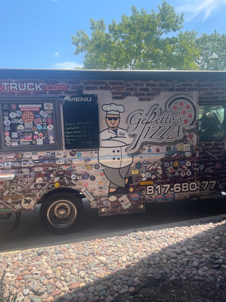 Side view of 'Gepetto's Pizza' food truck with a chef mascot graphic, adorned with various stickers, a visible menu board, and contact number, parked on a pebble-covered ground with trees in the background.