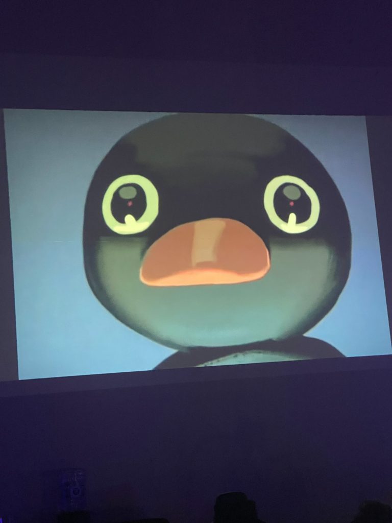 Close-up projection of a cartoon-like black duck's face with large eyes and an orange beak on a screen, in a dimly lit room.