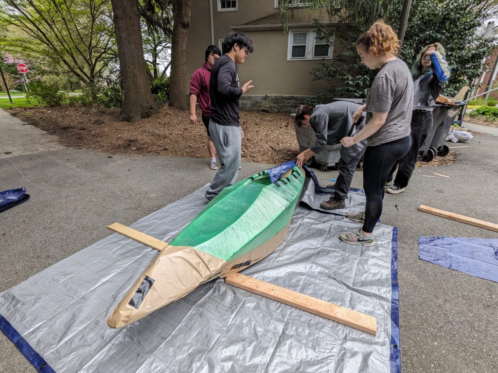several students crouching over a kayak-like boat wrapped in plastic and kraft paper
