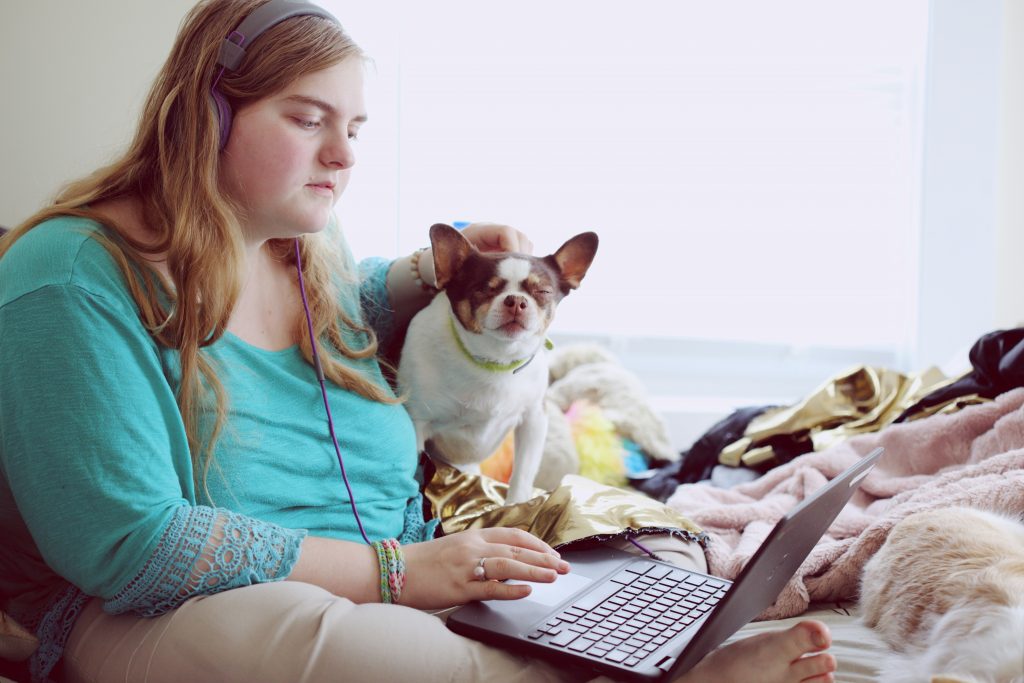 A young woman sits comfortably on a bed with her laptop, wearing headphones and focusing intently on the screen. Beside her, a small dog sits contentedly, enjoying a gentle pet. The cozy setting includes soft blankets and pillows, creating a relaxed and homey atmosphere. The natural light from a nearby window illuminates the scene, adding to the calm and peaceful environment.