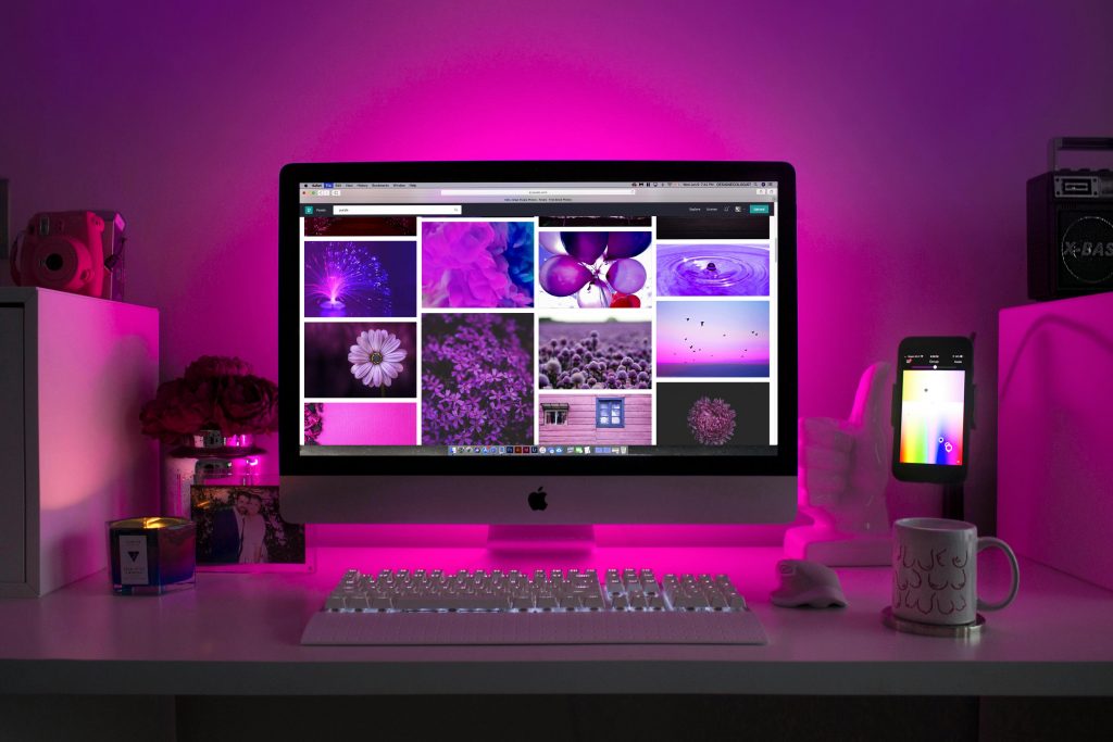 A desktop setup featuring an Apple computer displaying a grid of images in various shades of purple. The scene is bathed in a vibrant pink and purple glow, creating a cozy and artistic atmosphere. On the desk, there is a white keyboard, a white mouse, a smartphone on a stand displaying a color gradient, a coffee mug with a unique pattern, and decorative items including a candle, a jar of flowers, and a photo.