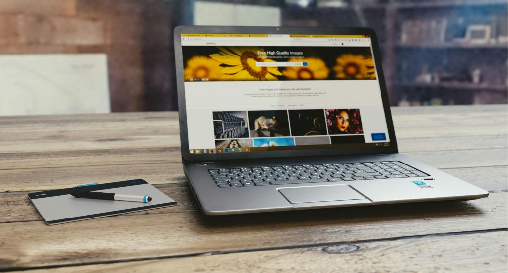 A sleek laptop sits on a wooden desk, displaying a website for free high-quality images. The screen shows a vibrant image of sunflowers and a gallery of various photos below. Next to the laptop is a digital drawing tablet with a stylus resting on top. The background is slightly blurred, indicating an indoor setting with shelves and decor items. The overall atmosphere is professional and creative, ideal for digital art or content creation.