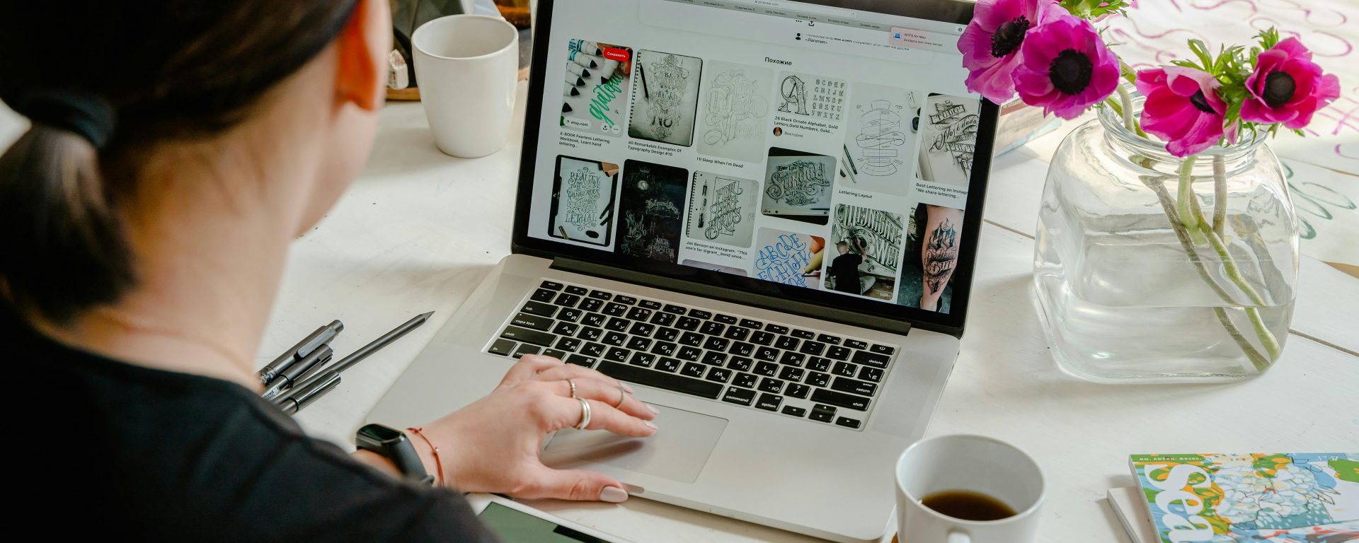 A person is working at a desk with a laptop open to a page of artistic sketches. They are holding a tablet displaying the word "creative" in stylized text. The desk is adorned with various items including a coffee mug, a book titled "Seasons," a vase with vibrant pink flowers, and writing utensils.