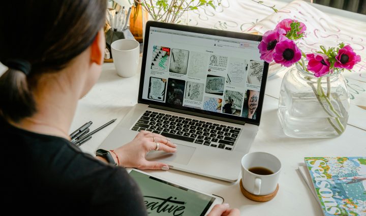 A person is working at a desk with a laptop open to a page of artistic sketches. They are holding a tablet displaying the word "creative" in stylized text. The desk is adorned with various items including a coffee mug, a book titled "Seasons," a vase with vibrant pink flowers, and writing utensils.