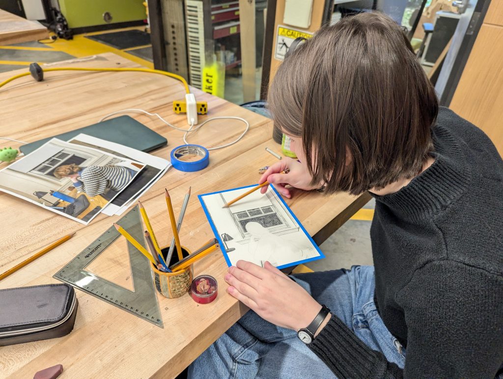 over-the-shoulder photograph of a student, sketching a scene from a photograph on a wood table