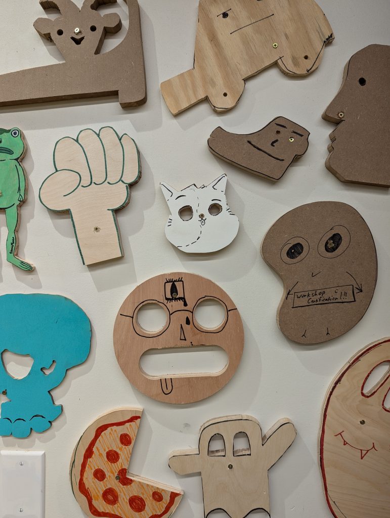 a group of small plywood shapes hanging on a flat wall, all carved into various amusing, monstrous faces and drawn on with markers and paint