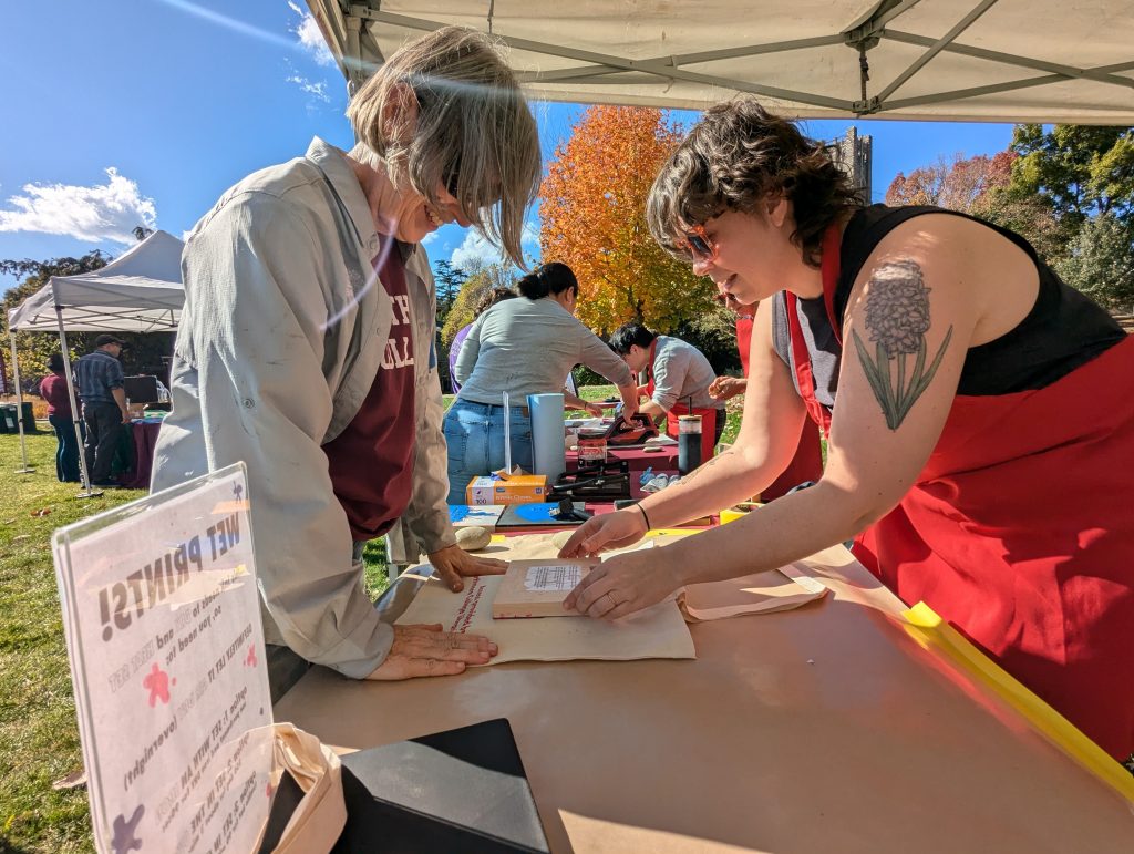 outdoor, table-level view of a group printing activity, with a swarthmore staff person in an apron helping someone with the project