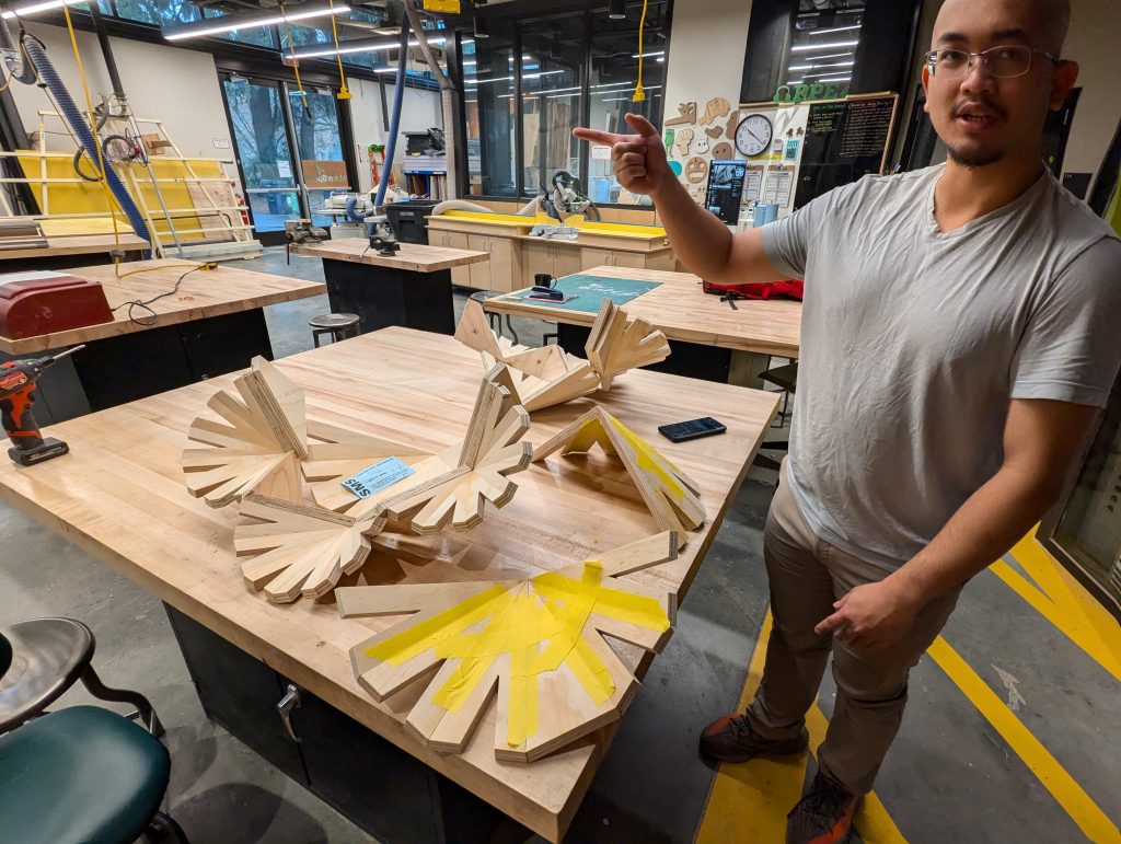 a student stands in front of his work on a woodshop table; the work is complex plywood shapes