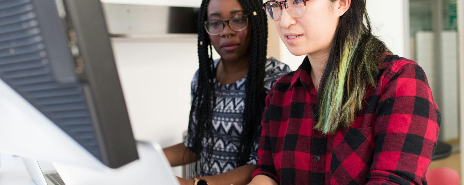 Two people collaborate at a computer workstation in a bright, modern office—one in a red-and-black plaid shirt and glasses types on a Mac while the other, wearing a patterned top with long braids, looks on attentively over her shoulder.