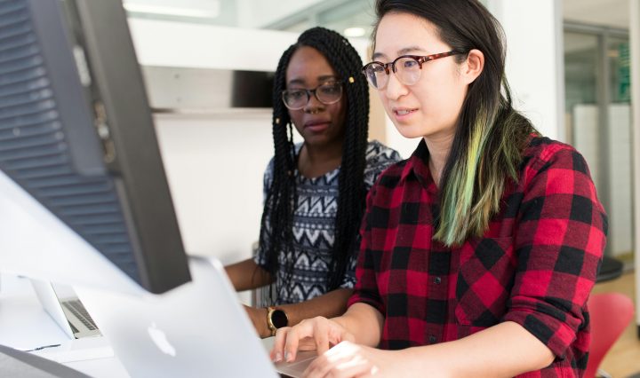 Two people collaborate at a computer workstation in a bright, modern office—one in a red-and-black plaid shirt and glasses types on a Mac while the other, wearing a patterned top with long braids, looks on attentively over her shoulder.