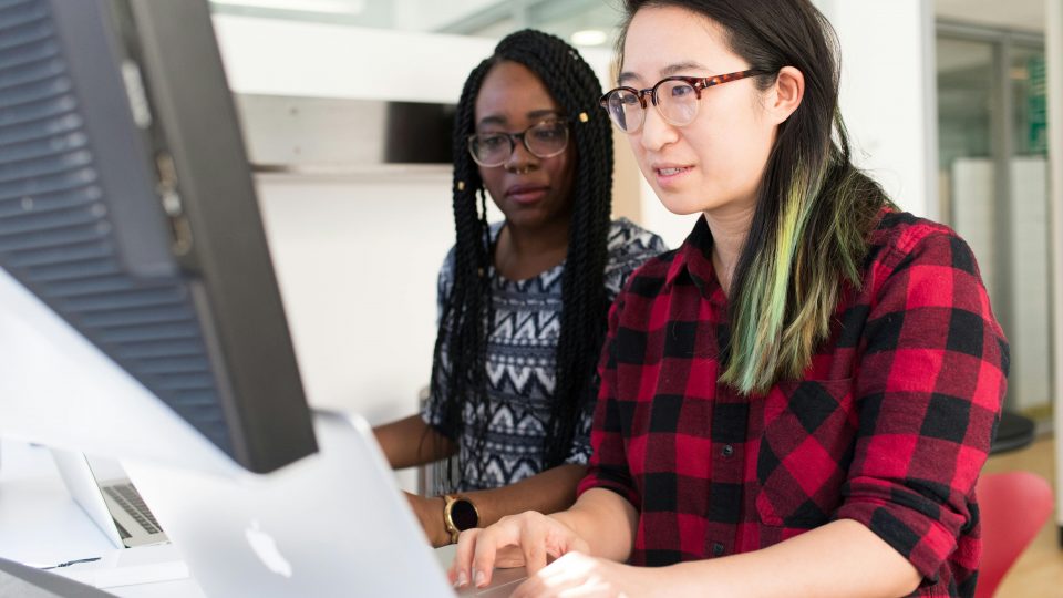 Two people collaborate at a computer workstation in a bright, modern office—one in a red-and-black plaid shirt and glasses types on a Mac while the other, wearing a patterned top with long braids, looks on attentively over her shoulder.