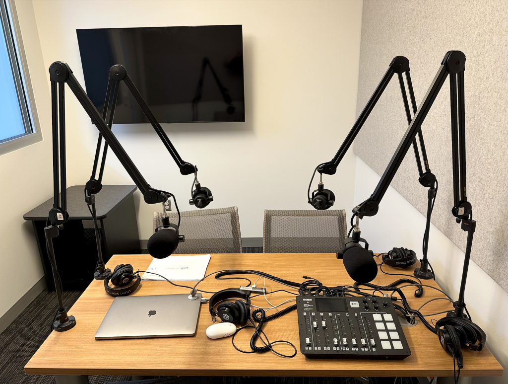 Table with audio mixer, 4 microphones on adjustable stands, 4 headsets and a laptop computer, a video display is mounted on the wall and soundproofing material is fixed to a wall.