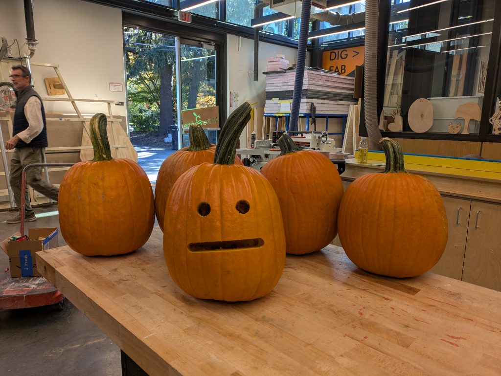 five pumpkins on a table, one in the foreground has a precisely carved emoji blank stare face carved