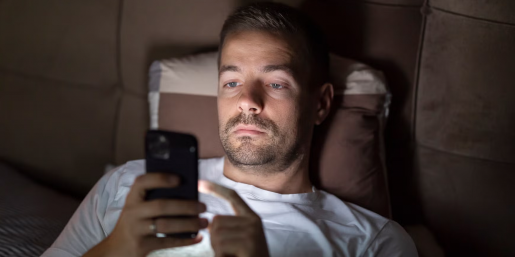 A man lays propped up in bed in a dark room scrolling through his cellphone.