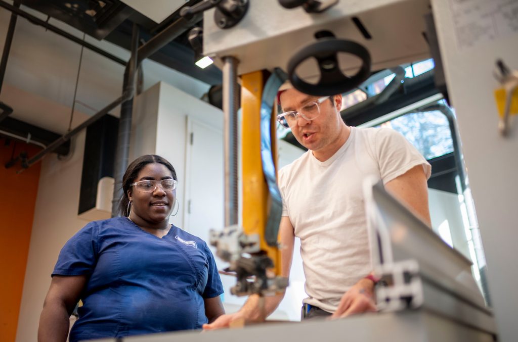 A makerspace staff member is photographed helping a workshop participant on a bandsaw, shot through the tool in the foreground