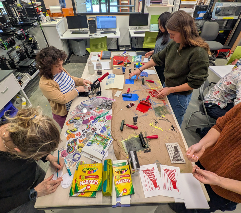 half a dozen students and staff working around a table covered in art and printmaking supplies