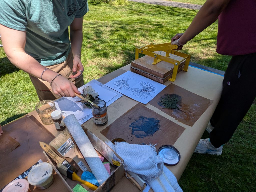 an outdoor scene of two students making prints from natural foliage. The table has a small hand printing press, several finished prints, and various bottles of pigment and brushes