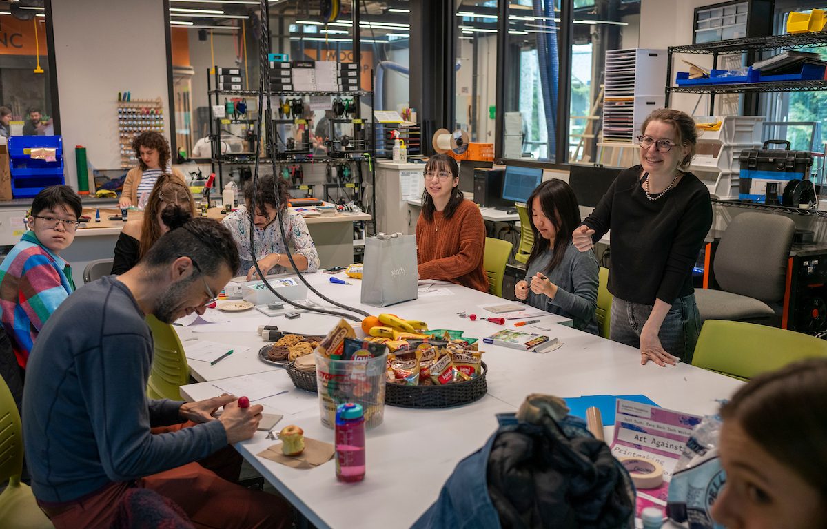 Students participate in an “Art Against AI” stamp-making workshop in Swarthmore College’s MakerSpace, gathered around worktables with art supplies and refreshments as facilitator Linda Huber leads the activity.